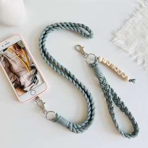 May include: A light blue macrame phone lanyard with a silver-tone clasp and a wooden bead charm spelling "LAUREN". The lanyard is attached to a pink phone case with a phone inside.