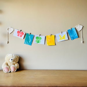 Op de afbeelding: Een reeks kleurrijke kunstwerken hangt aan een muur met een teddybeer die op een houten tafel eronder zit. De kunstwerken zijn gemaakt met vingerverf en bevatten een rode bloem, een groene klaver, een gele vlinder en een blauw abstract ontwerp.