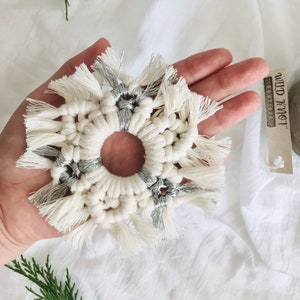 Peut inclure: Une d&eacute;coration de No&euml;l en macram&eacute; en forme de flocon de neige blanc et argent avec des franges. L'ornement est tenu dans une main sur un fond blanc.
