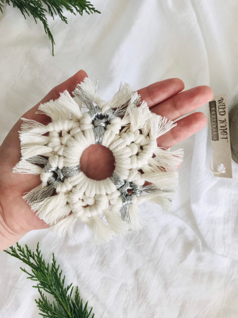 Peut inclure: Une d&eacute;coration de No&euml;l en macram&eacute; en forme de flocon de neige blanc et argent avec des franges. L'ornement est tenu dans une main sur un fond blanc.
