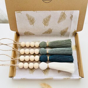 Peut inclure: Un ensemble de quatre pompons décoratifs dans une boîte en carton. Chaque pompon est composé de perles en bois et d'une touffe de fils dans les tons de vert et de blanc. La boîte est doublée de papier à motifs de fougères.