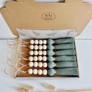 Può includere: Un set di otto ornamenti con perline di legno e nappe in una scatola di cartone. Le perline sono in legno naturale e le nappe sono di un verde salvia spento. La scatola è foderata con carta a motivi e ha un logo che dice "Wild Knot".