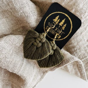 May include: A pair of green tassel earrings with gold hooks. The earrings are on a black card with the text "Wild Knot" and a logo of three pine trees.