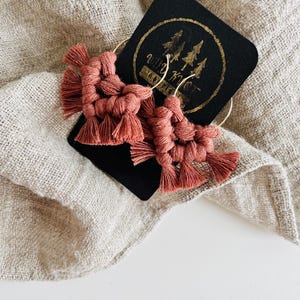 May include: A pair of terracotta-coloured macrame earrings with gold hoops. The earrings are handmade and feature a tassel design. The earrings are displayed on a black card with the text "Wild Knot Co."
