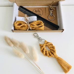 May include: A craft kit in a cardboard box includes a black comb, measuring tape, and mustard-colored cord. A macrame keychain with a tassel is displayed next to the box. The kit also includes a keychain clip and a card with the text "Wild Knot".