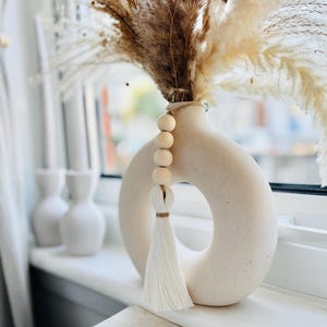 May include: A cream-colored, donut-shaped vase with a tassel and wooden bead accent, filled with dried pampas grass and other dried plants. The vase sits on a white windowsill, with two other white vases in the background.
