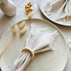 May include: A dining table arrangement with speckled ceramic plates, linen napkins with fringed edges, and wooden napkin rings. The scene is enhanced with dried bunny tail grass and a decorative tassel. A pink wine glass is also visible, creating a rustic and elegant aesthetic.