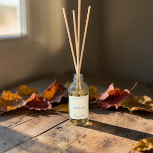 May include: A clear glass reed diffuser bottle filled with light yellow liquid, featuring a white label with the text "Anchored Candle Co." and "Chai Latte." Four light-coloured wooden reeds are inserted into the bottle, extending upwards.