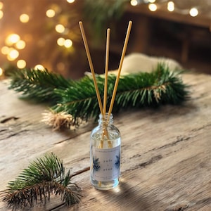 May include: A clear glass aromatherapy diffuser bottle with three wooden reeds. The label reads "Yule + Pine". The bottle sits on a wooden surface with pine branches and blurred lights in the background. The overall theme is festive decor.