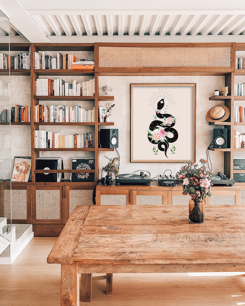 May include: A black and white illustration of a snake with flowers and stars on a beige background. The illustration is framed and hanging on a wall in a room with a wooden table and shelves.