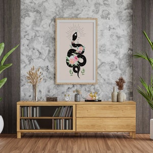 May include: A framed print of a black snake with pink and white flowers on a pink background. The snake is coiled and has a star on its head. The print is hanging on a wall with a wooden shelf below it.