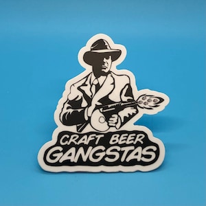Op de afbeelding: Zwart-witte sticker van een persoon met een fedora en een pak die een pistool vasthoudt met een bierhopontwerp. De tekst "CRAFT BEER GANGSTAS" staat onder de persoon.