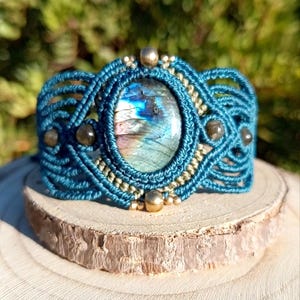 May include: A teal macrame bracelet featuring a central oval labradorite gemstone with blue and gold iridescence. The woven cord design is accented with small metallic beads and four smaller dark round stones arranged symmetrically around the centre.