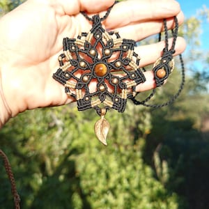 May include: A macrame pendant necklace featuring a black and tan cord design in a geometric star pattern. The center contains a round brown tiger eye gemstone, accented with small metallic beads and a dangling bronze-toned leaf charm.
