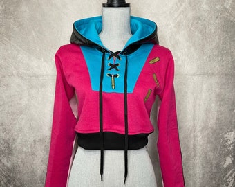 May include: A pink and blue cropped hoodie with black trim and a lace-up detail at the front. The hoodie has a black hood and features three gold bullet-shaped embellishments on the right side. The text "La Couturière de Paris" and "FAUBOURG SAINT-HONORÉ" is printed on the bottom of the hoodie.