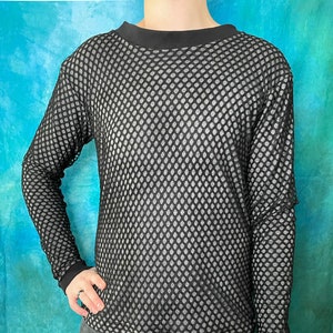 Könnte beinhalten: Schwarzes Mesh-Langarmshirt mit Rundhalsausschnitt und schwarzen Bündchen. Das Shirt hat ein rautenförmiges Muster und wird mit einer dunkelgrauen Hose kombiniert. Der Hintergrund ist ein Türkis-Blau-Verlauf.