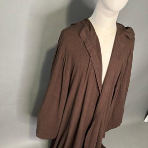Peut inclure: Une robe à capuche marron avec des manches longues. La robe est faite d'un tissu doux et fluide et a un devant large et ouvert. La capuche est attachée à la robe et est conçue pour être portée sur la tête. La robe est exposée sur un mannequin.