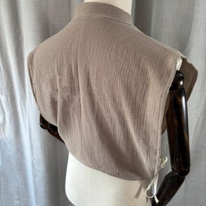May include: A tan-colored, sleeveless garment with a high collar, displayed on a mannequin. The fabric has a textured, crinkled appearance. White ties are visible on the sides. The garment appears to be a top or accessory.