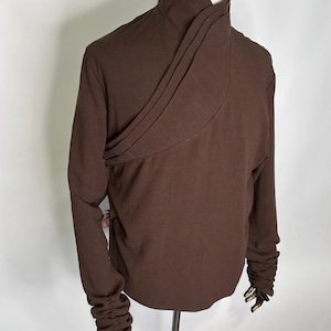 May include: A long-sleeved brown shirt featuring a high, wrap-style collar with layered pleats. The garment has a solid dark brown color and includes gathered, ruched detailing along the lower portion of both sleeves, extending to the cuffs.