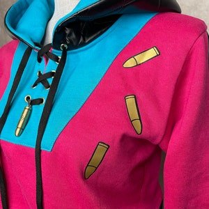 Pu&ograve; includere: Una felpa con cappuccio corta con cappuccio in pelle nera, blocchi di colore blu e rosa e grafiche di proiettili dorati.