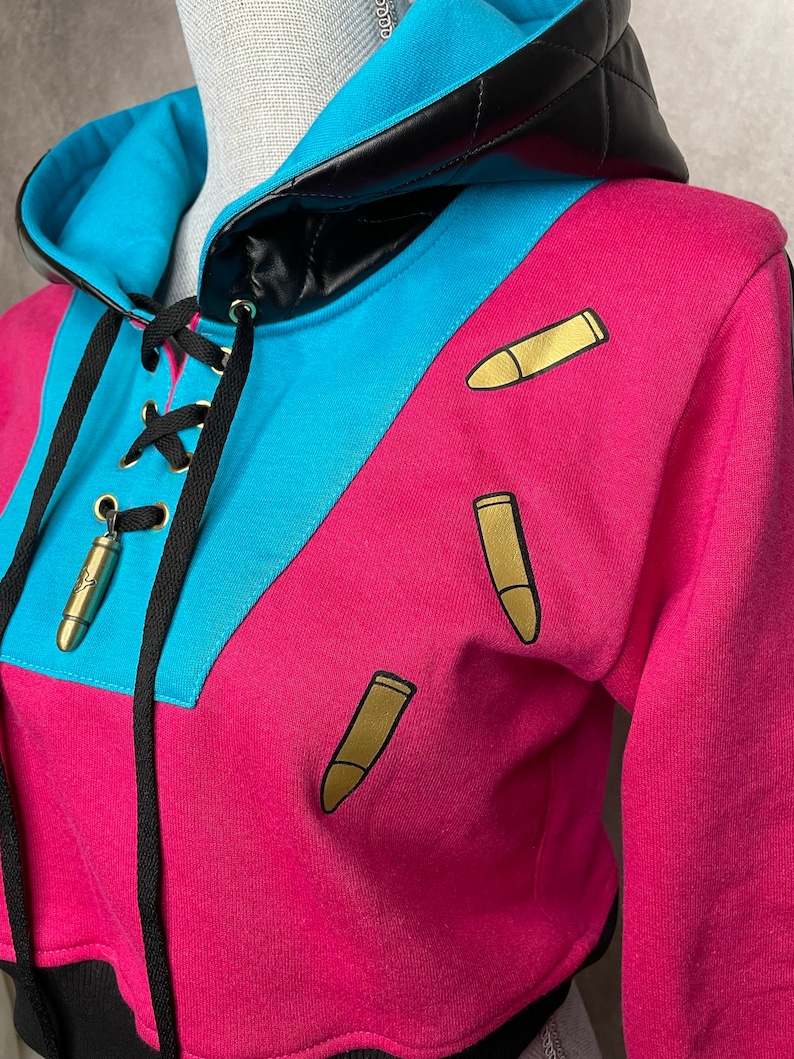 Pu&ograve; includere: Una felpa con cappuccio corta con cappuccio in pelle nera, blocchi di colore blu e rosa e grafiche di proiettili dorati.