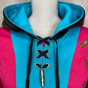 Pu&ograve; includere: Una felpa rosa e blu con un dettaglio di pizzo nero e un ciondolo a forma di proiettile dorato. La felpa ha un bordo in pelle nera intorno al cappuccio.