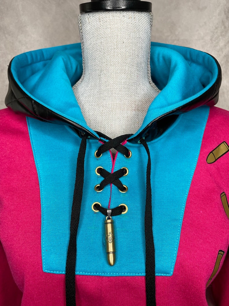 Pu&ograve; includere: Una felpa rosa e blu con un dettaglio di pizzo nero e un ciondolo a forma di proiettile dorato. La felpa ha un bordo in pelle nera intorno al cappuccio.