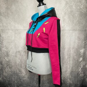 Pu&ograve; includere: Una felpa con cappuccio corta rosa, blu e nera con una striscia bianca e nera sulla manica. La felpa ha un cappuccio con coulisse e ciondoli a forma di proiettile dorati.