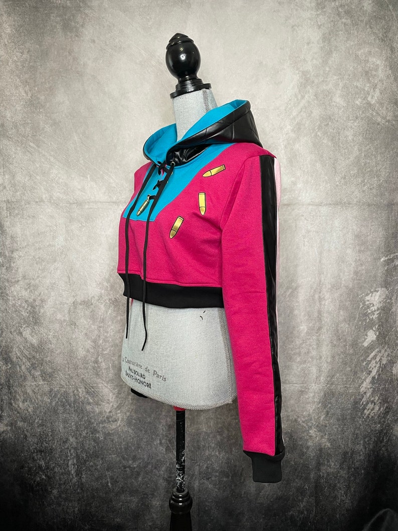 Pu&ograve; includere: Una felpa con cappuccio corta rosa, blu e nera con una striscia bianca e nera sulla manica. La felpa ha un cappuccio con coulisse e ciondoli a forma di proiettile dorati.