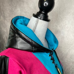 Pu&ograve; includere: Una felpa rosa e blu con un cappuccio trapuntato in pelle nera e un dettaglio di pizzo. La felpa ha un ciondolo a forma di proiettile dorato attaccato al pizzo.