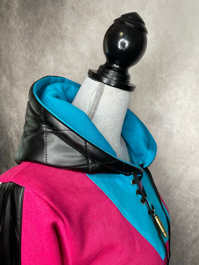 Pu&ograve; includere: Una felpa rosa e blu con un cappuccio trapuntato in pelle nera e un dettaglio di pizzo. La felpa ha un ciondolo a forma di proiettile dorato attaccato al pizzo.