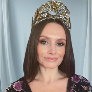May include: A woman wearing a gold-coloured crown decorated with clear crystals and ornate details. The crown rests on long, dark brown hair. The woman is wearing a black top with a floral design.