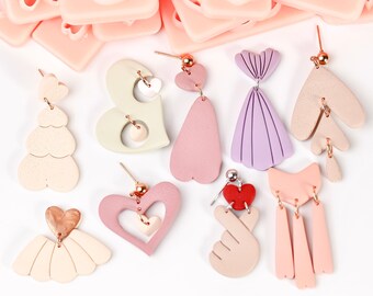 Puede incluir: Surtido de pendientes de arcilla polimérica en varias formas y colores, incluyendo corazones, un vestido, un árbol y un gesto de mano. Los pendientes están hechos a mano y presentan una variedad de texturas y detalles.