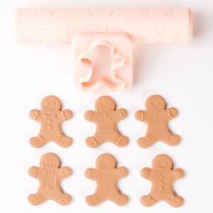Op de afbeelding: Een set met peperkoekman-koekjesvormen en een deegroller met een peperkoekman-ontwerp. De vormen zijn roze en de deegroller is roze met een wit ontwerp. Er zijn zes peperkoekmannen op een wit oppervlak.