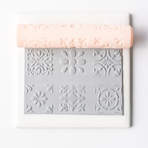 May include: A pink rolling pin with a repeating pattern of floral designs. The rolling pin is on top of a gray square tile with six different embossed patterns.