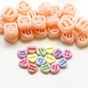 May include: A collection of pink heart-shaped candy molds with various words inscribed on them, such as "You Suck", "Not Yours", "No Way", and "Hard Pass".  The molds are arranged in a semi-circle around a group of colorful heart-shaped candies with different messages.