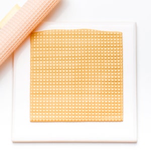 May include: A close-up of a waffle cookie sheet, with a waffle iron roller in the background. The cookie sheet is white with a light brown waffle pattern.