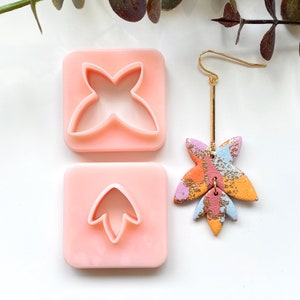Puede incluir: Dos cortadores de arcilla polimérica rosa, uno con forma de trébol de cuatro hojas y el otro con forma de lágrima. Un par de pendientes hechos de arcilla polimérica con un diseño floral en tonos rosa, naranja y azul.