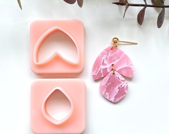 May include: Two pink plastic molds for making earrings, one shaped like a teardrop and the other like a curved leaf. A pair of pink marbled earrings with gold accents are shown next to the molds.