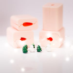 Puede incluir: Un conjunto de cinco adornos navideños en miniatura, que incluyen dos árboles de Navidad verdes y tres muñecos de nieve blancos. Los adornos están dispuestos sobre una superficie blanca con un fondo borroso de moldes de silicona rosa.