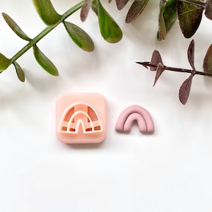 Op de afbeelding: Een roze plastic koekjesvormpje in de vorm van een regenboog en een roze kleiregenboog. De koekjesvormpje zit in een vierkant frame.