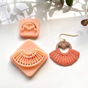 Peut inclure: Trois moules en argile polymère rose pour la fabrication de boucles d'oreilles. Les moules sont en forme d'éventail, de cercle avec découpe et de demi-cercle. Une paire de boucles d'oreilles en pompons de couleur pêche est également présentée.