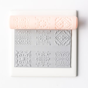 May include: A set of six silicone embossing stamps with intricate geometric designs. The stamps are arranged in a 2x3 grid on a white surface. A pink silicone rolling pin is shown above the stamps.