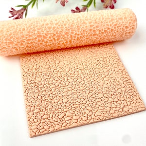 Op de afbeelding: Een beige getextureerde deegroller en een bijpassend vierkant vel. De deegroller heeft een herhalend patroon van onregelmatige vormen. Het vel heeft hetzelfde patroon, waardoor een craquelé-effect ontstaat. De items zijn waarschijnlijk voor knutselen of bakken.