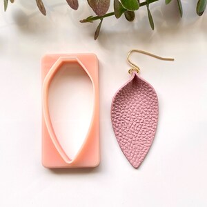 Puede incluir: Un pendiente de cuero rosa con forma de hoja y un gancho dorado. También se muestra un molde de plástico rosa, que se utiliza para crear el pendiente.