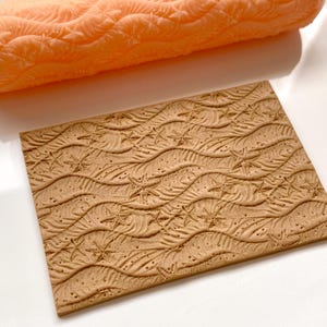 Pode incluir: Um rolo de massa castanho com um desenho em relevo de ondas e estrelas do mar. O rolo é usado para criar um padrão em relevo em fondant ou argila.