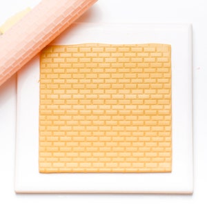 May include: A white plastic rolling pin with a yellow handle and a brick pattern rolling surface. The rolling pin is on top of a white plastic sheet with a brick pattern embossed on it.