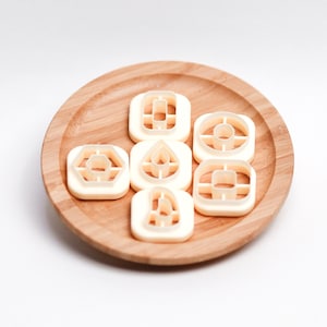 May include: A set of seven white plastic cookie cutters on a wooden plate. The cutters are shaped like a diamond, a circle, a teardrop, a rectangle, a hexagon, a square, and a leaf.