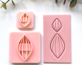 May include: Three pink polymer clay stamps with a geometric design. The stamps are in the shape of a diamond with a series of lines inside. The stamps are on a white surface.