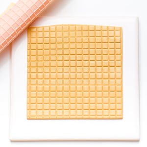 May include: A white square baking mat with a grid pattern in a light brown color. The mat is on a white surface.